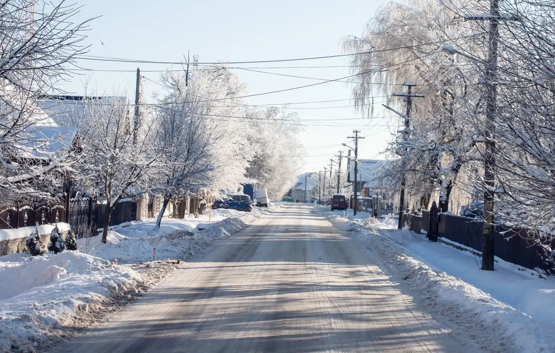 A serene winter scene of a snow-covered road lined with leafless trees and houses under a clear blue sky.