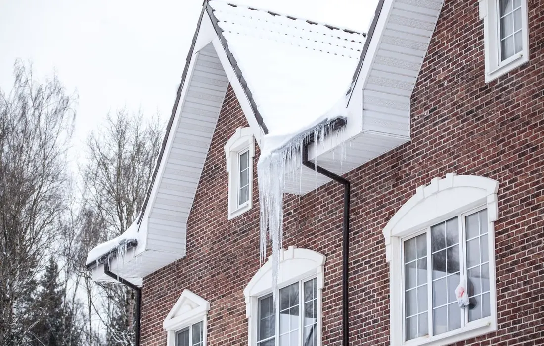 A brick house with snow-covered roof and icicles in a winter setting.