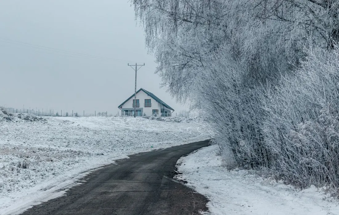 A serene winter scene featuring a snow-covered rural house and frosty trees along a winding road.