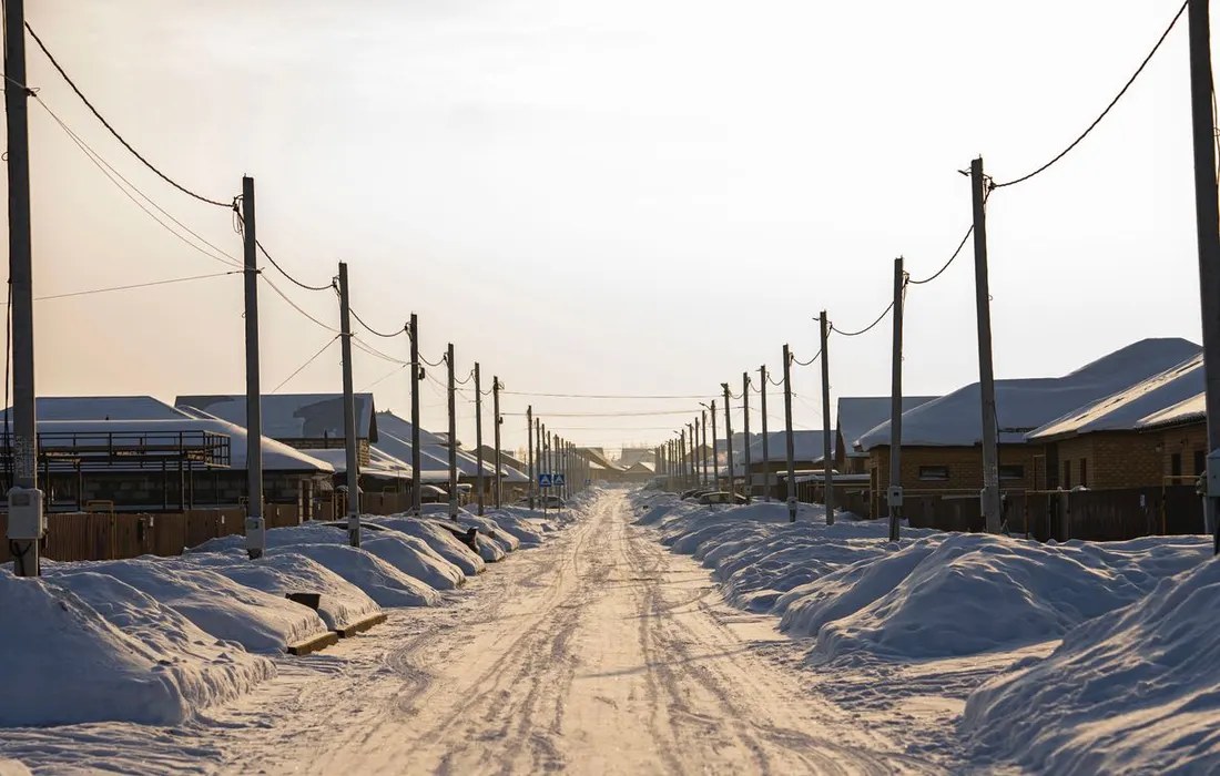 A tranquil snow-covered road lined with houses and power lines under a soft sunset in winter.