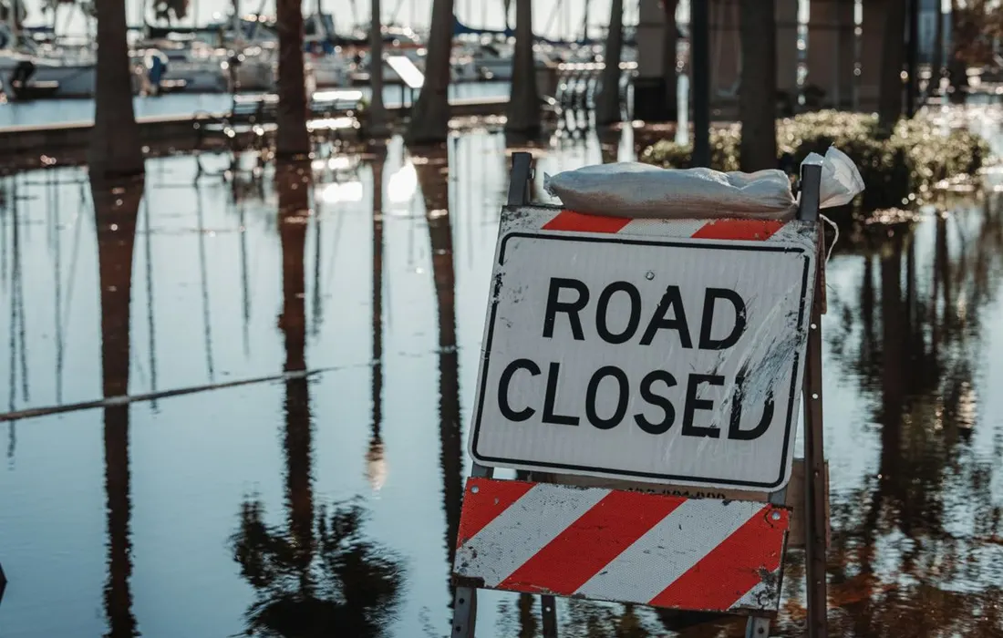 Road closed sign amidst flooded street, reflecting calm water and trees.