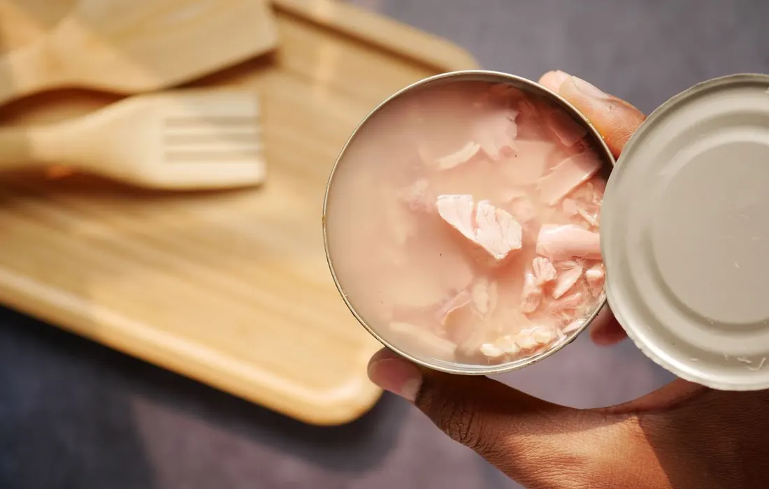 Hands holding an opened tin of tuna over a chopping board with kitchen utensils.