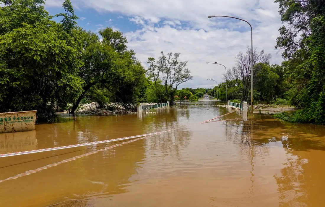 A flooded road with a submerged bridge, surrounded by trees, under a cloudy sky.