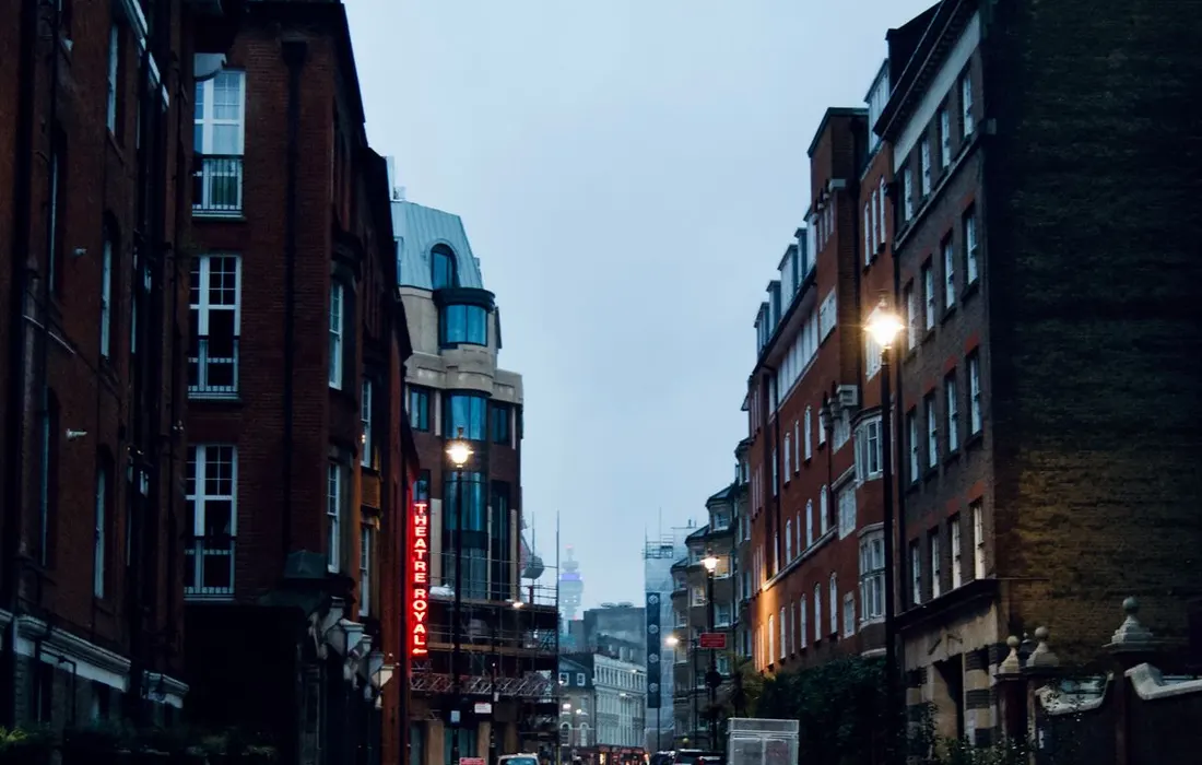 A wet city street lined with tall buildings and illuminated signs on a rainy day.