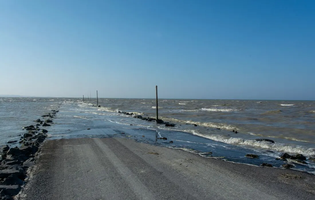 A flooded coastal road surrounded by the sea and horizon under a clear blue sky.