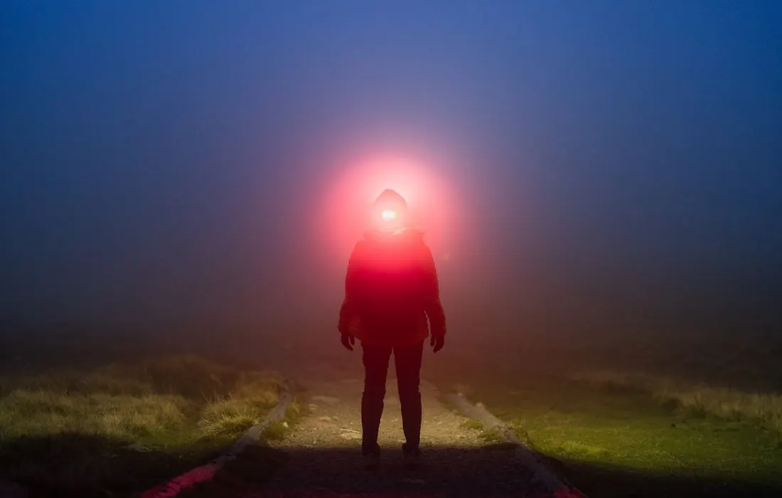 A person using a headlamp on a foggy outdoor path at night.