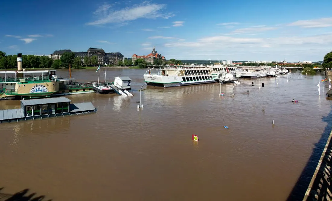 Partially submerged boats on a flooded river under a clear blue sky in cityscape setting.