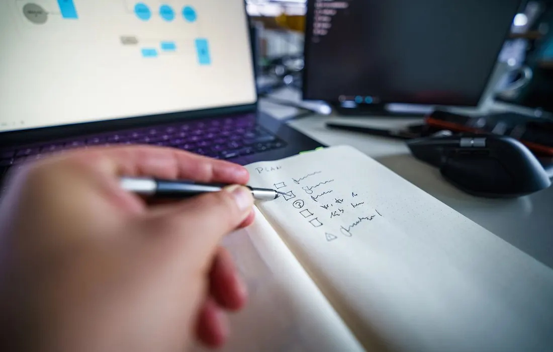 A hand writing in a notebook beside a laptop.