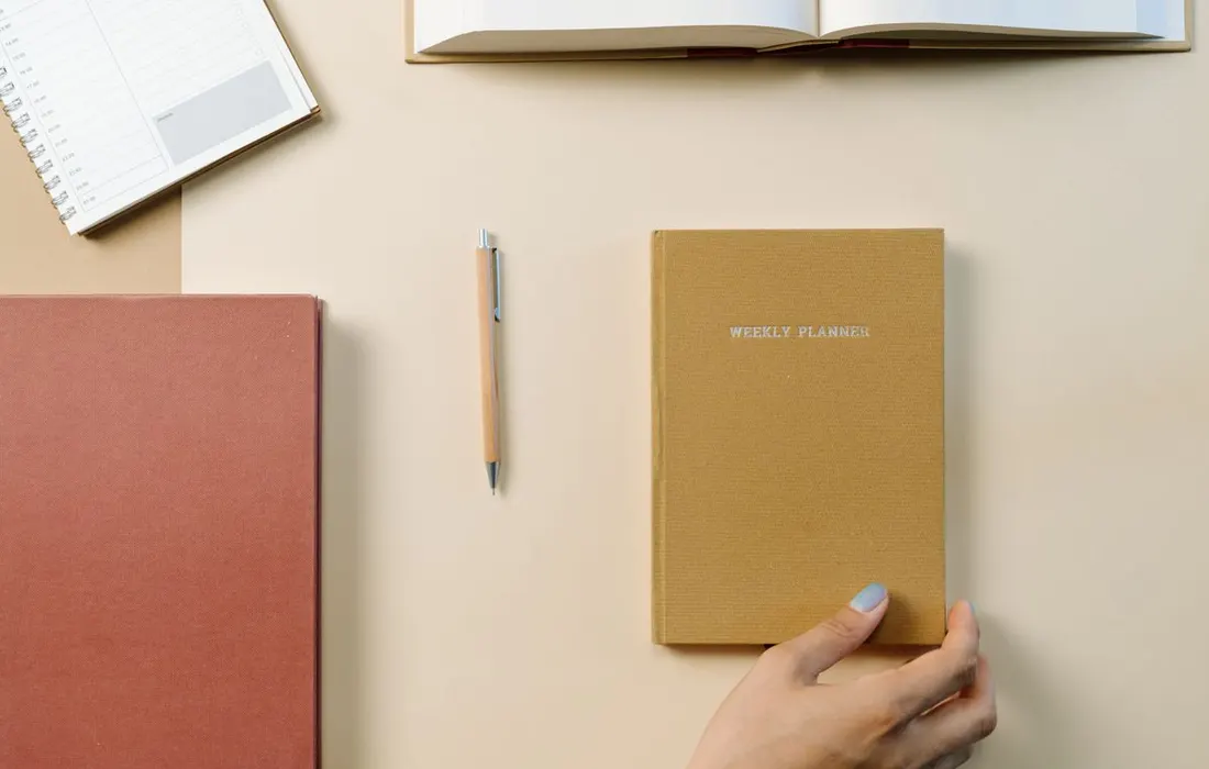 An open notebook, weekly planner and pen arranged on a desk.
