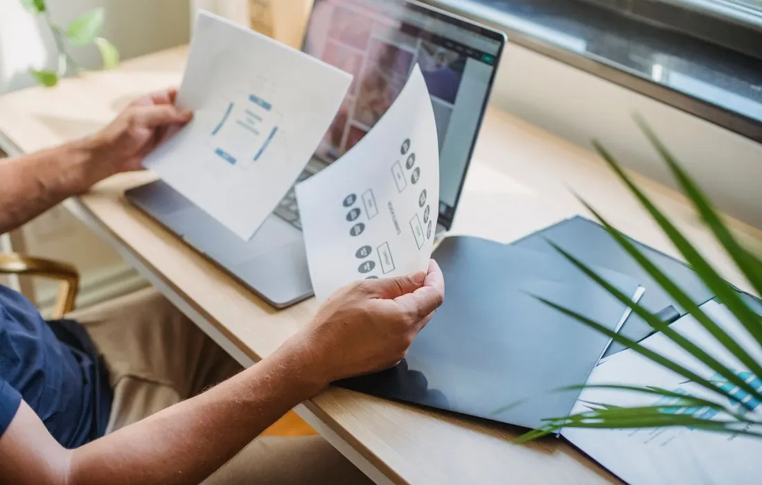 Anonymous hands reviewing printed papers beside a laptop.