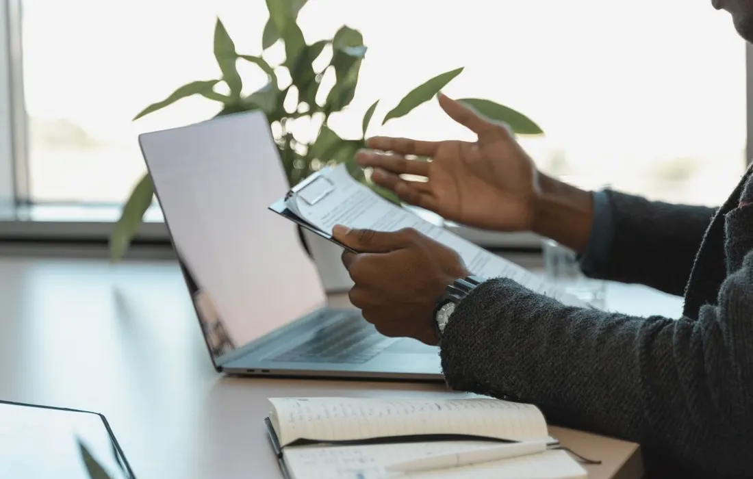 Hands holding printed documents beside a laptop and notebook.
