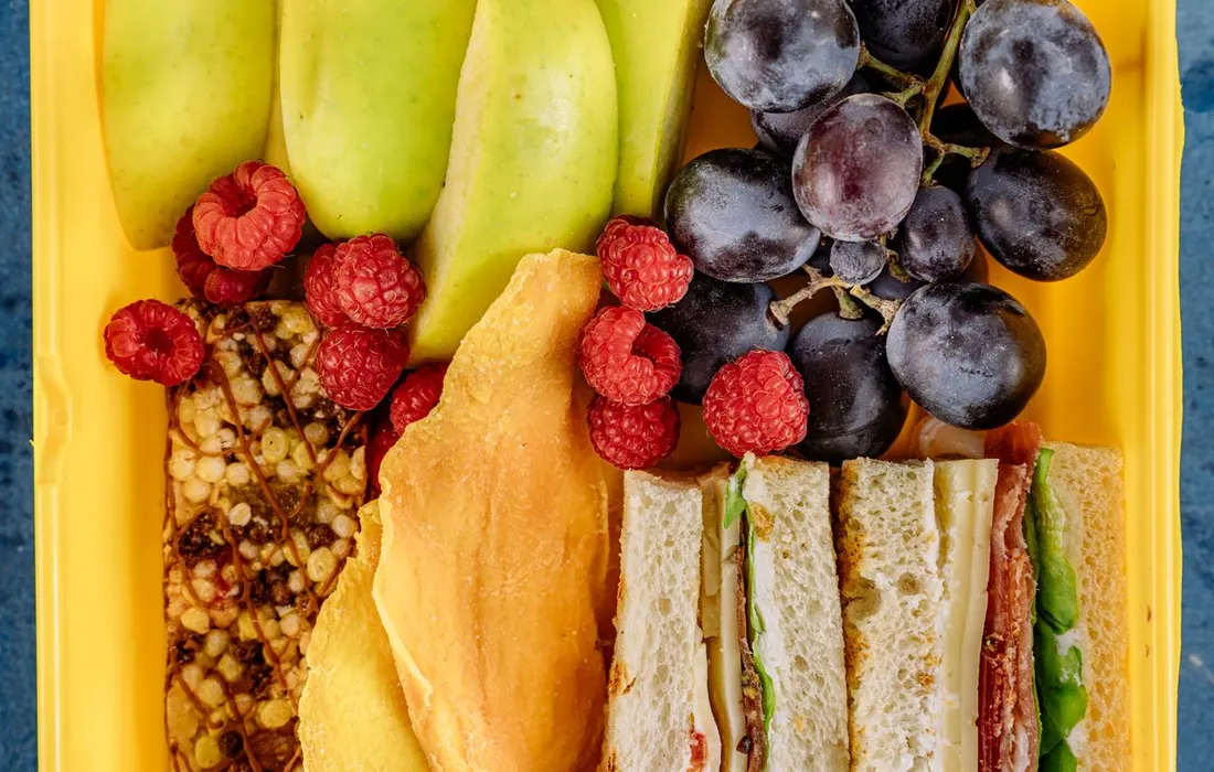 A lunchbox containing a sandwich, fruit and a snack bar.