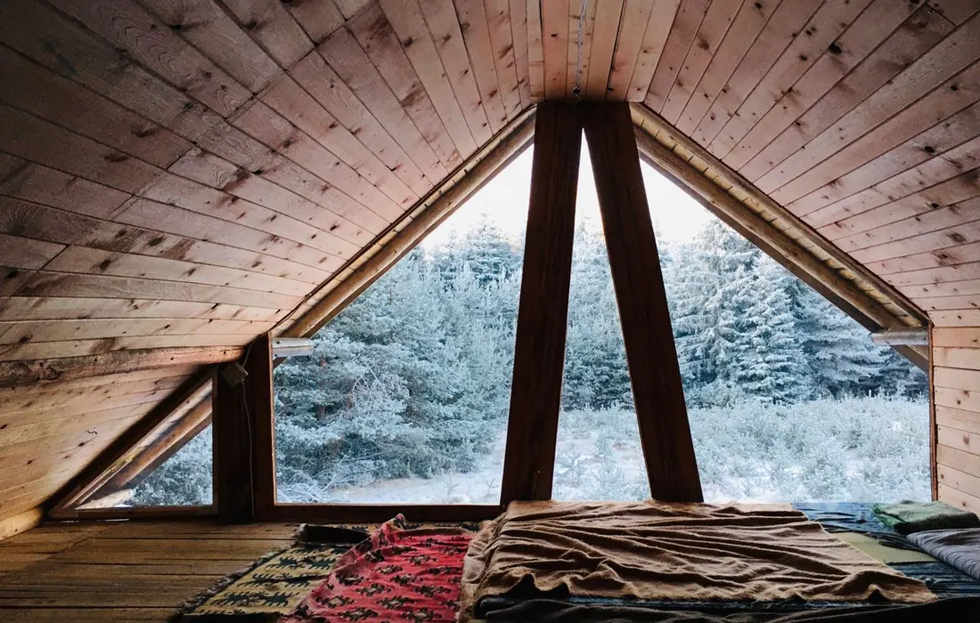 Warm attic room in wooden cabin with blankets overlooking a snowy forest.