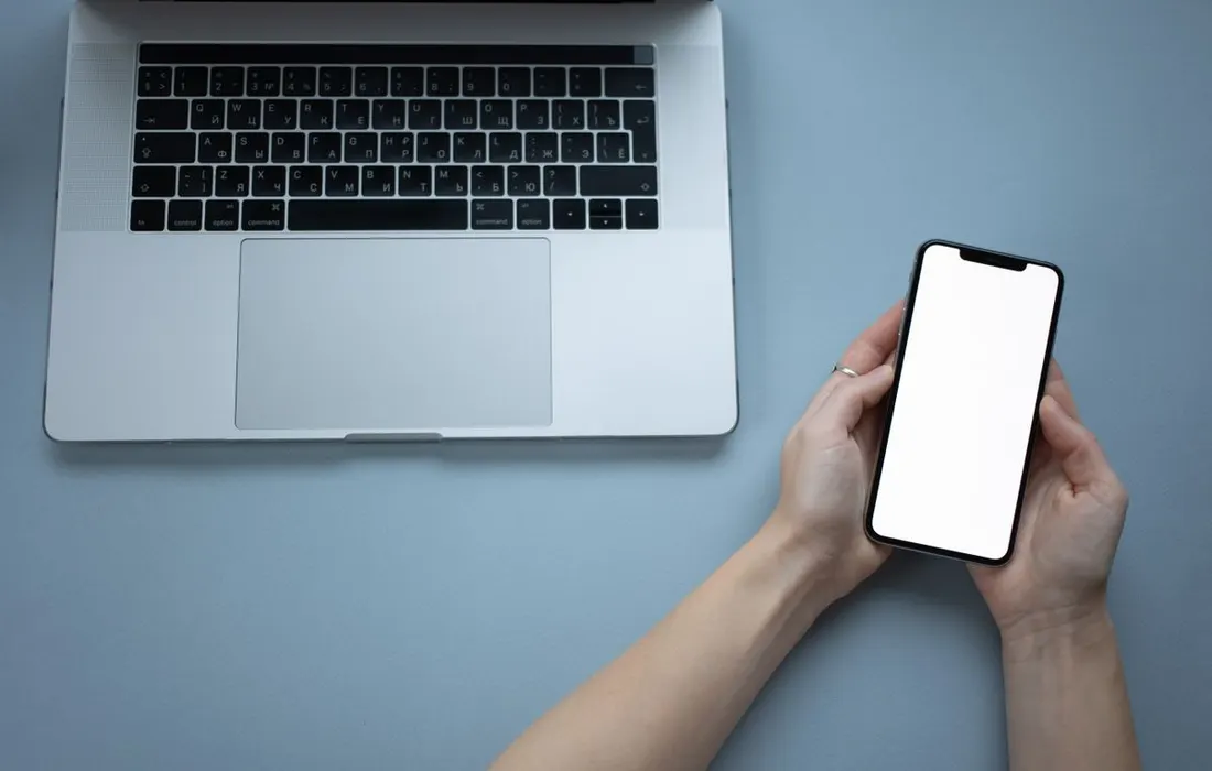 Hands holding a smartphone beside a laptop.
