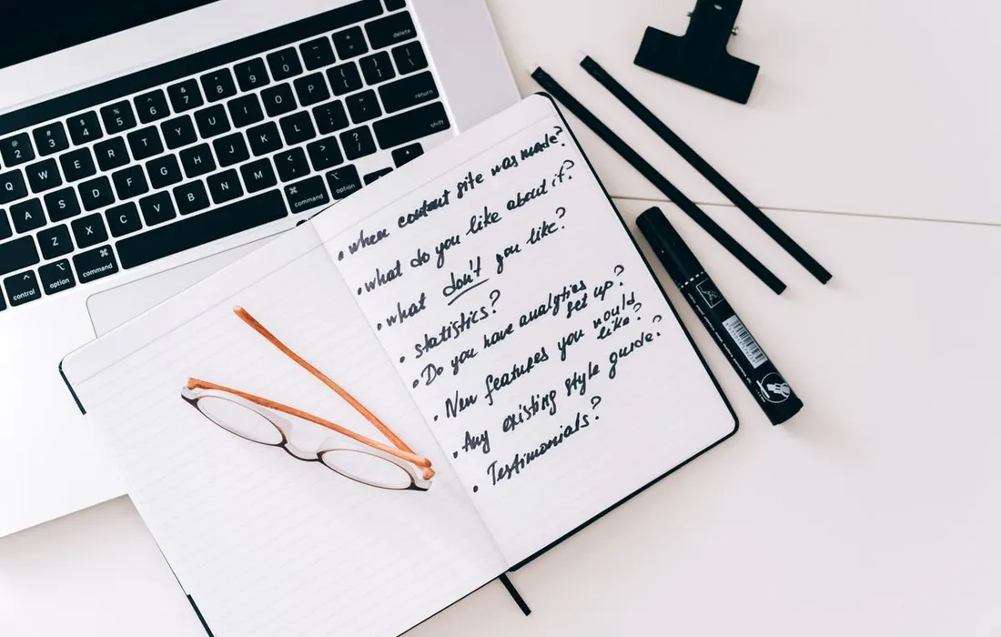 An open notebook, glasses and a laptop on a desk.